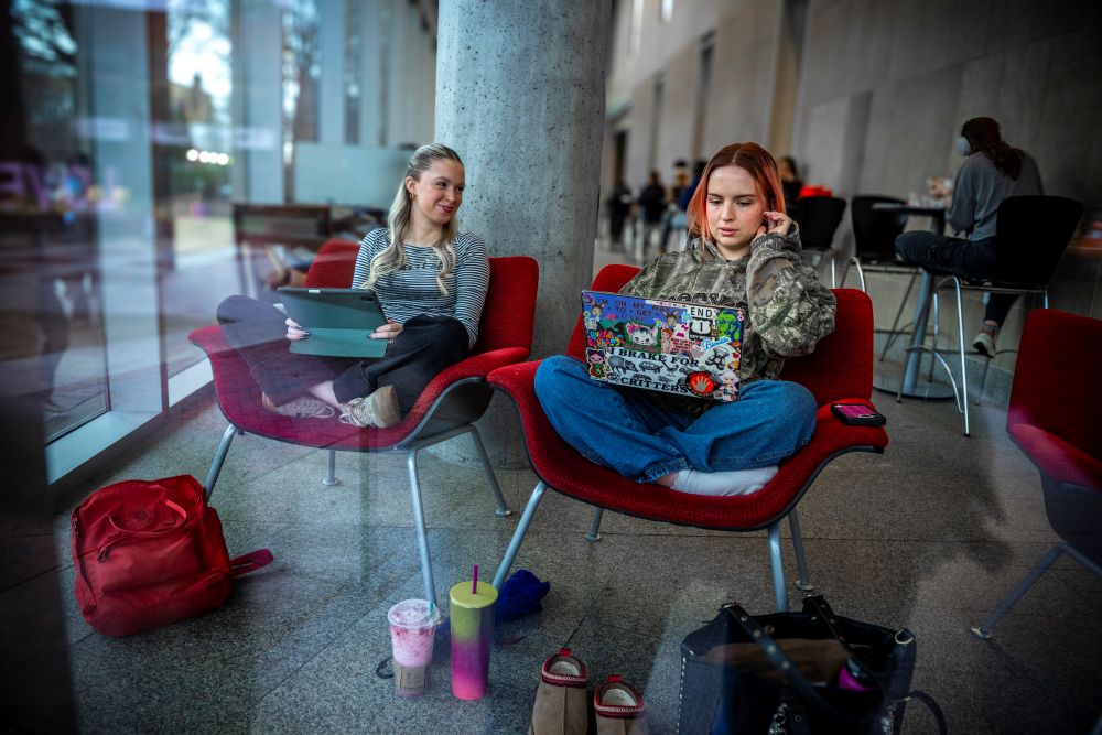 Two female students sitting in red foldout chairs near the entrance of James Cabell Branch Library, working on their laptops