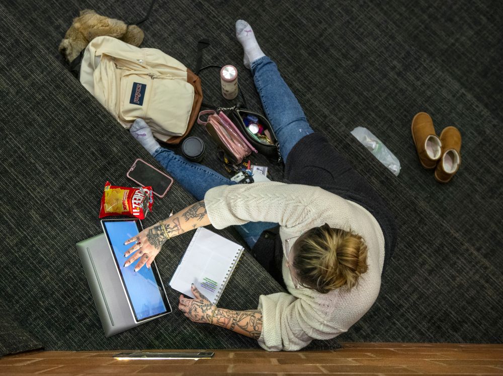 An overhead shot of a student sitting on steps working on their laptop, surrounded by school-related clutter