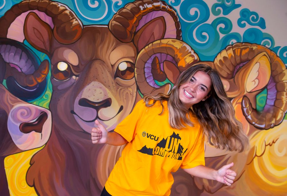 Female student wearing VCU UNcommon t-shirt posing with thumbs up next to a mural of Rodney the Ram